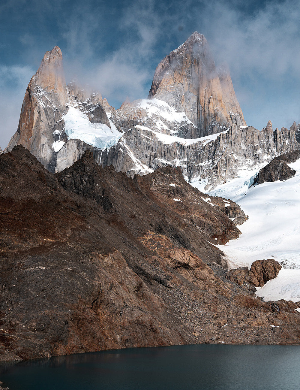 Lake with jagged, cloud and snow covered mountains behind it