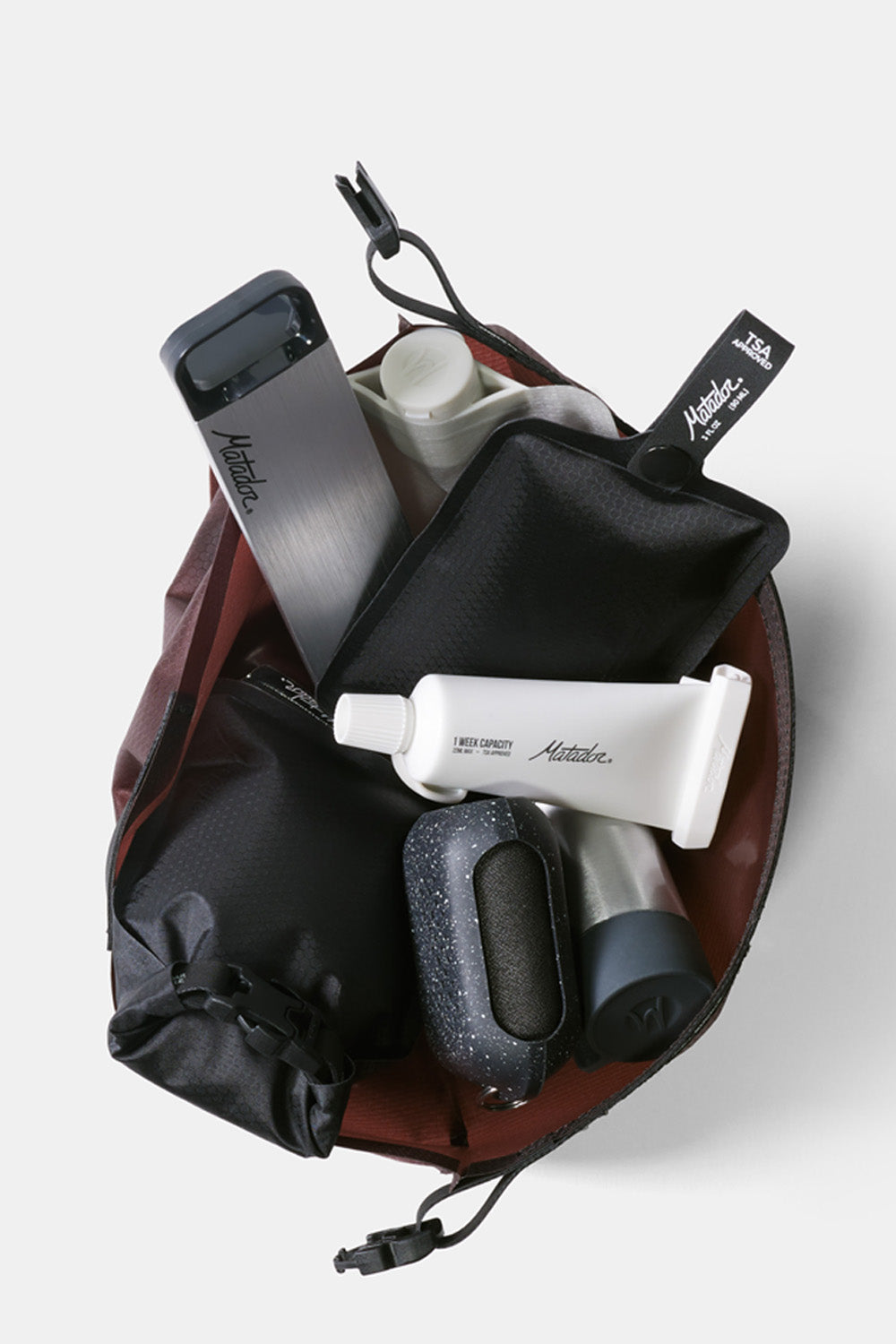 top view of garnet toiletry case full of various matador toiletry items like pill canister, toothpaste tube, soap bar case, and trek towel.