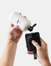 Person pouring liquid from a white bottle with a black nozzle into a black pouch on a light gray background.