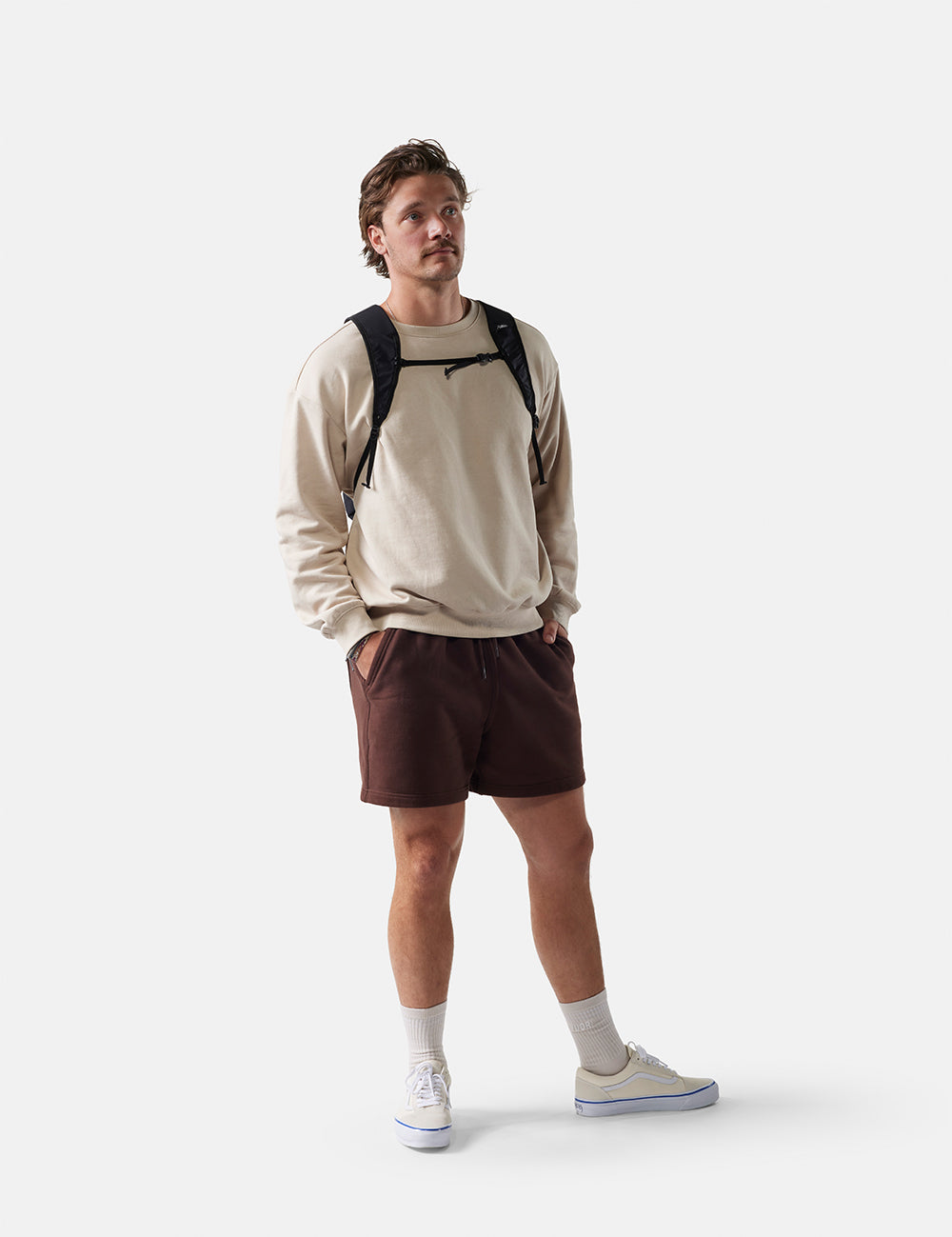 Man wearing a beige sweatshirt, brown shorts, and white sneakers on a white background