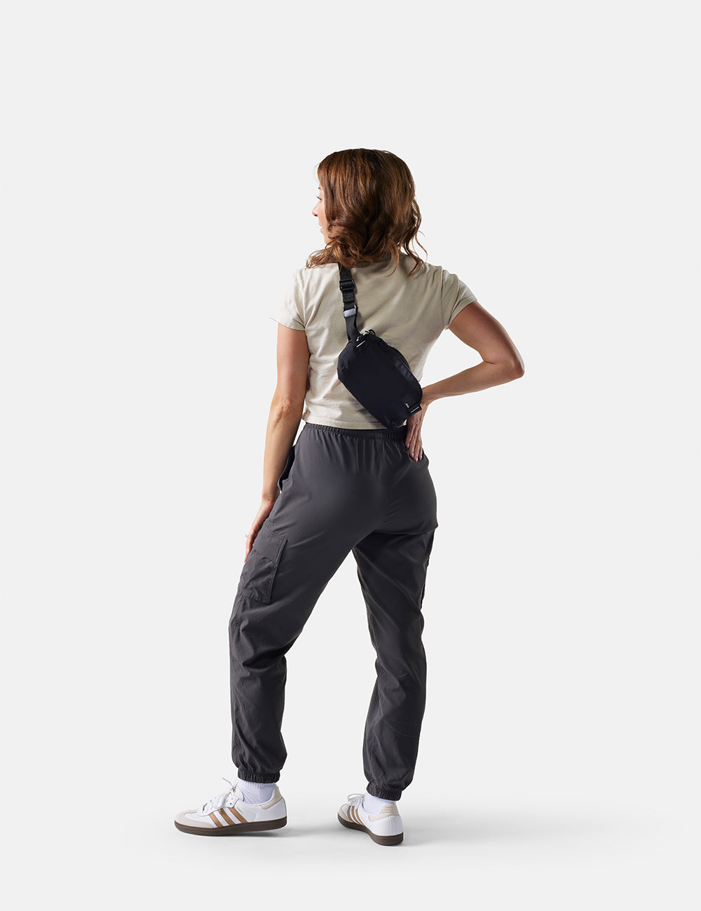 Person wearing dark gray pants and a beige shirt with a crossbody bag on a white background