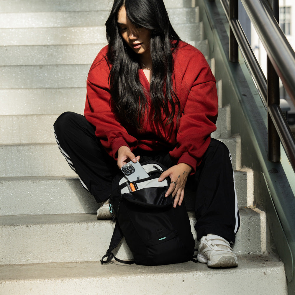 woman in read sweatshirt, sitting on stairs placing phone in her black backpack