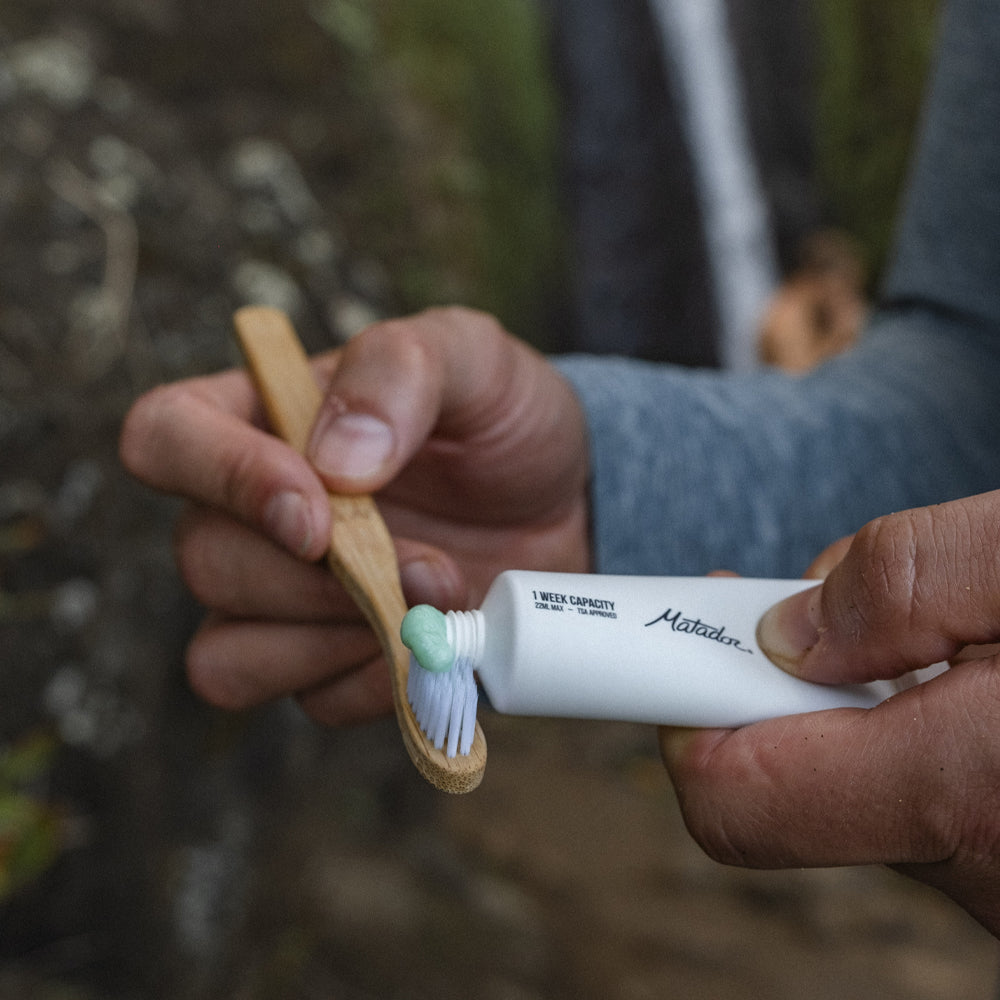person near waterfall squeezing toothpaste onto bamboo toothbrush